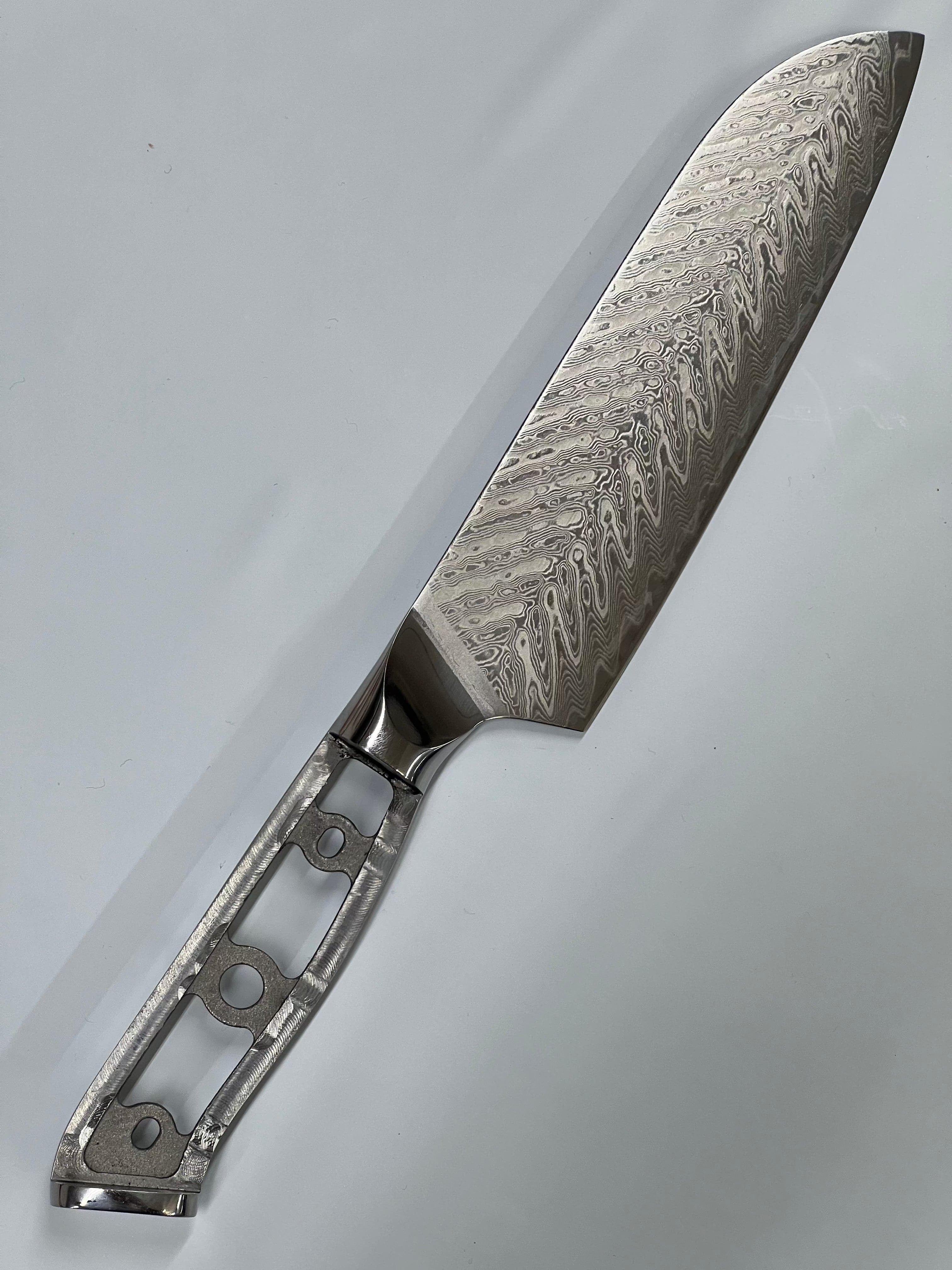 A Sild Santoku - VG10 Damask Kokkekniv 7" med et rustfri damask stålblad med intrikate bølgemønstre. VG10 eggstål bladet er høypolert, og håndtaket er åpen ramme med utskjæringer for redusert vekt. Bakgrunnen er en vanlig, lys grå overflate .